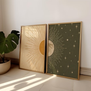 May include: Two framed art prints. One shows a sunburst design in yellow and gold on a cream background. The other depicts a moon and stars in gold on a dark green background. Both are in wooden frames.
