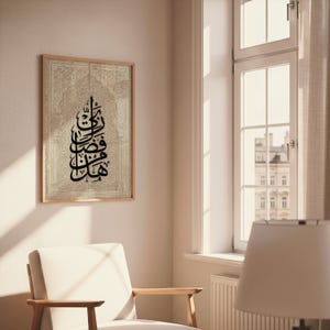 May include: A framed artwork featuring Arabic calligraphy in black against a beige background with an ornate, textured design. The artwork is displayed on a wall in a room with a window and a chair.