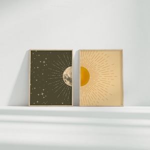 Two unframed poster prints, one dark olive green featuring stars and a crescent moon and the other pale yellow with a sun illustration, standing side-by-side on a white surface against a white wall.