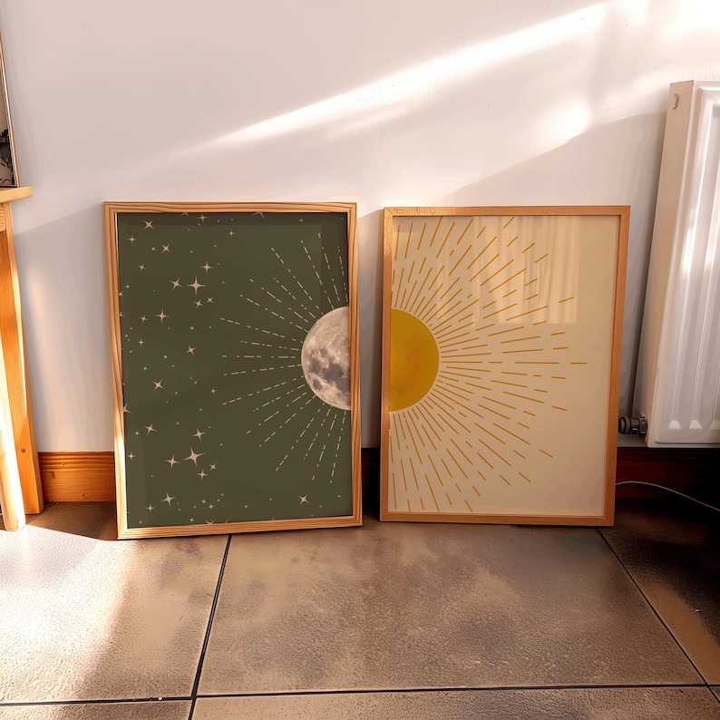 Two framed celestial art prints leaning against a white wall on a tiled floor beneath sunlight, featuring a dark green moon print on the left and a yellow sun print on the right.