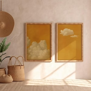 May include: Two framed prints with cloud designs are displayed on a wall. The artwork has a yellow backdrop with white clouds. A woven hat hangs above, and a potted plant and woven baskets sit below, creating a warm, inviting aesthetic.