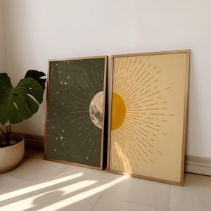 Two framed posters leaning against a white wall next to a large potted monstera plant, featuring olive green moon art and yellow sun art.