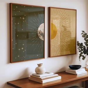 Framed green moon art and yellow sun art hanging on a white wall above a long wooden console table holding stacked books and a ceramic vase.