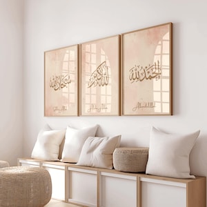 Puede incluir: Un conjunto de tres láminas de arte mural rectangulares en marcos de madera clara, con fondos de acuarela beige, caligrafía árabe en marrón oscuro y las transliteraciones al inglés correspondientes: Subhan Allah, Allahu Akbar y Alhamdulillah.