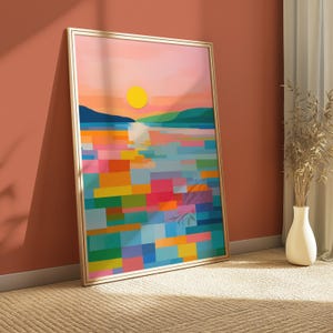 Puede incluir: Una impresión artística abstracta enmarcada que representa una puesta de sol sobre el agua. La obra de arte utiliza un estilo geométrico con bloques de color en tonos de rosa, naranja, amarillo, azul y verde. El marco es dorado.