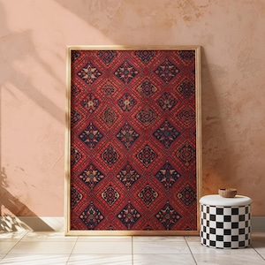 May include: A framed, rectangular rug with a detailed, repeating pattern in red, navy blue, and beige. The rug is in a gold-colored frame, leaning against a peach-colored wall. A black and white checkered stool with a wooden bowl sits nearby.
