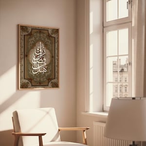 Könnte beinhalten: Ein gerahmtes Kunstwerk mit kunstvoller arabischer Kalligraphie in Weiß auf einem gemusterten grün-braunen Hintergrund. Der Rahmen ist hellbraun. Das Kunstwerk hängt an einer weißen Wand in der Nähe eines Fensters.