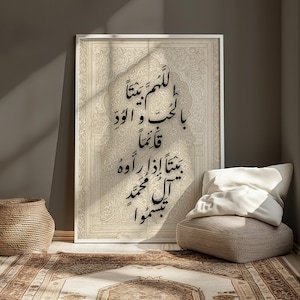 May include: A framed print featuring Arabic calligraphy in black script on a beige background. The print is leaning against a wall, with a woven basket and a patterned rug in the foreground. A beige cushion and pillow are on the right.