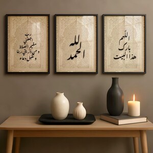 May include: Three framed art prints with Arabic calligraphy above a wooden table. The prints have a beige background and black frames. On the table are white vases, a black vase, a candle, and a book. The table is made of light-coloured wood.