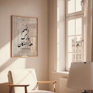 Puede incluir: Una impresión enmarcada con un intrincado patrón de color claro y caligrafía árabe en negro. El texto dice "O Allah, bendice este lugar". La obra de arte se exhibe en una pared junto a una ventana.