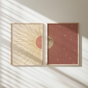 May include: Two framed art prints. One shows a sunburst design with a peach circle on a cream background. The other print has a moon with radiating lines and stars on a reddish-brown background. Both are in light wood frames.