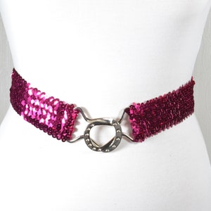 Op de afbeelding: Een roze paillettenriem met een zilveren gesp. De riem is elastisch en heeft een simpel ontwerp.