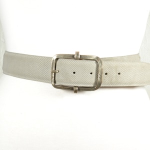 May include: A light gray belt with a large, square, silver-toned buckle. The belt has a perforated texture and is displayed on a white mannequin. The buckle has a rectangular shape with a central bar and two smaller bars on top.