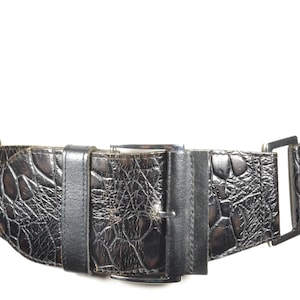 May include: A wide, black belt with a crocodile-skin texture. The belt features a large buckle and a series of smaller, rectangular metal accents. The belt is designed to be worn around the waist and is a fashion accessory.