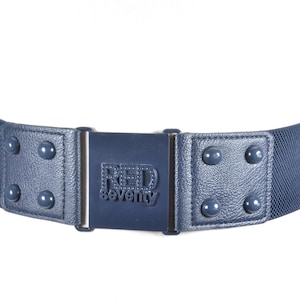 Può includere: Una cintura elastica blu navy con una fibbia argentata e una placca rettangolare blu navy con il testo "RED seventy" in lettere bianche. La cintura ha una superficie testurizzata ed è decorata con borchie argentate.