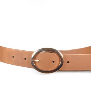 May include: A tan leather belt with a gold-colored oval buckle. The belt is shown on a white mannequin. The belt has three holes for adjustment.