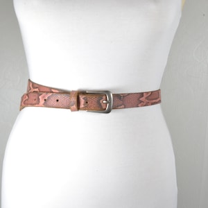 Può includere: Una cintura in pelle rosa con motivo a stampa di pelle di serpente e fibbia argentata.