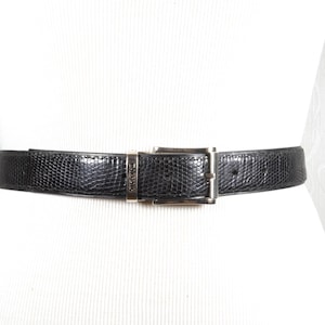Op de afbeelding: Een zwarte leren riem met een zilveren gesp. De riem heeft een getextureerd, gegraveerd patroon.