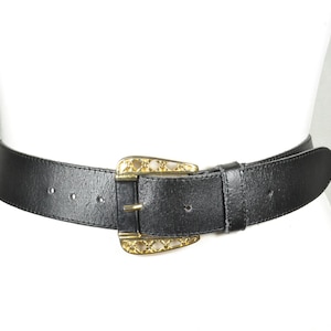 May include: A black leather belt with a decorative gold-colored buckle. The belt is wide and has a textured surface. The buckle has an ornate design. The belt is displayed on a white mannequin.