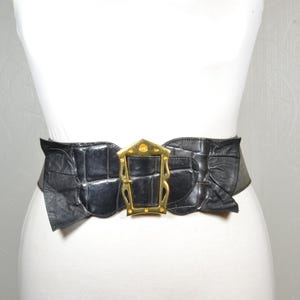 Puede incluir: Un cinturón ancho de color negro con un acabado texturizado de imitación de cocodrilo y una hebilla de metal dorado prominente. La hebilla tiene un marco rectangular con recortes decorativos y un perno central, fijado al material negro fruncido.