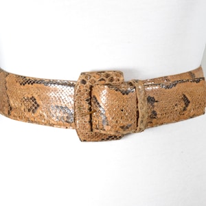 Può includere: Una cintura in pelle di serpente marrone con una grande fibbia rettangolare. La cintura è realizzata in vera pelle e ha un design classico e senza tempo.