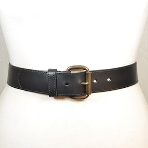 Pode incluir: Um cinto largo de couro preto com acabamento liso e uma fivela retangular de metal em tom de latão antigo. O cinto inclui um passador de couro preto a condizer e costuras visíveis ao longo das bordas, fechado com um pino único.