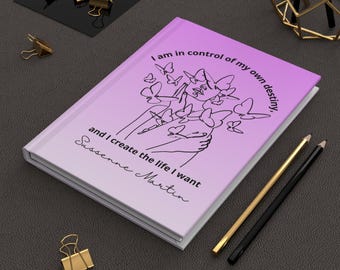 May include: A hardcover journal with a gradient pink and purple cover. The cover features a line drawing of a person holding butterflies, with the text "I am in control of my own destiny, and I create the life I want, Sussenne Martin."