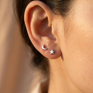 Peut inclure: Gros plan sur une oreille ornée de deux petites boucles d'oreilles argentées. L'une est en forme de croissant de lune, l'autre en forme d'étoile à cinq branches. Les boucles d'oreilles sont simples et élégantes.