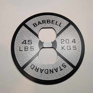 May include: A black barbell weight plate with a silver textured surface. The plate is labeled "BARBELL STANDARD" and has the weight "45 LBS" and "20.4 KGS" printed on it.