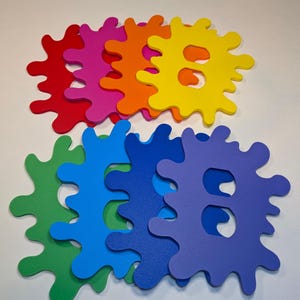 May include: A set of eight colorful foam shapes, each resembling a paint splatter. The shapes are in the colors of the rainbow, with red, orange, yellow, green, blue, indigo, and violet. Each shape has a cutout in the center, forming a letter 'B'.