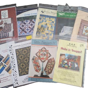 Könnte beinhalten: Eine Sammlung von Steppmustern in transparenten Plastikhüllen. Die Muster zeigen verschiedene Quilt-Designs, darunter geometrische Muster, Blumenmotive und Landschaftsbilder. Einige Muster haben Text wie "Criss Cross" und "Make it Snappy!"