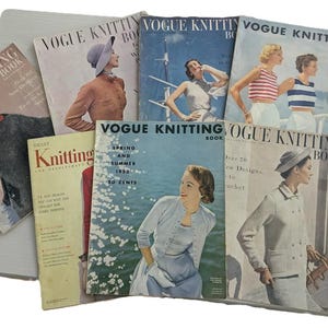 Op de afbeelding: Een verzameling vintage Vogue Knitting Book-tijdschriften, met kleurrijke covers met illustraties van vrouwen in stijlvolle breikleding. De tijdschriften tonen retro mode en design uit het midden van de 20e eeuw, met tekst als "Spring and Summer 1953".
