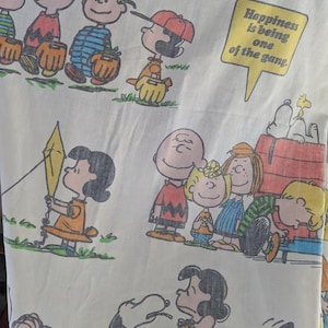 May include: A white fabric with a Peanuts theme. The fabric features various Peanuts characters, including Charlie Brown, Snoopy, Lucy, and Linus. The fabric also includes a speech bubble with the text "Happiness is being one of the gang!"
