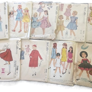 May include: A collection of vintage McCall's sewing patterns for children's clothing. The patterns feature illustrations of dresses, skirts, and jumpsuits. The covers display colorful drawings of the finished garments, with text indicating sizes and styles.