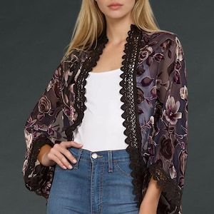 Peut inclure: Un kimono floral transparent dans les tons de violet, gris et noir. Le kimono présente une bordure en dentelle noire le long des bords et des manches. Le vêtement est porté sur un haut blanc et un jean bleu.