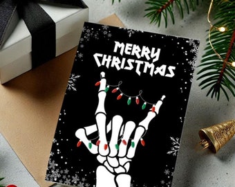 May include: A white gift box with a black ribbon sits next to a Christmas card. The card features a black background with a skeleton hand making a rock and roll gesture, strung with Christmas lights. The words "MERRY CHRISTMAS" are printed at the top.