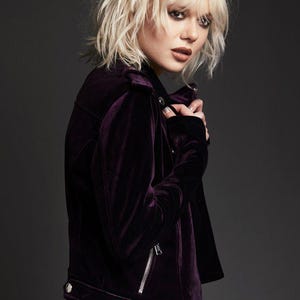 May include: A dark purple velvet jacket with a silver zipper and silver buttons. The jacket has a classic biker style with a notched collar and a tailored fit. The model is wearing a white shirt underneath.