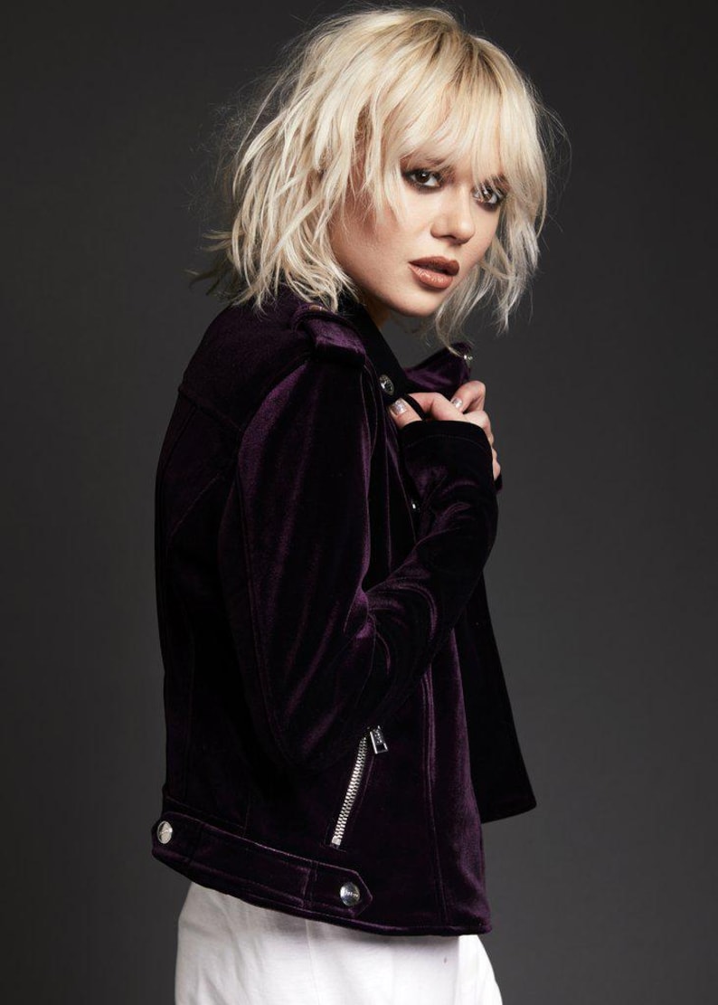 May include: A dark purple velvet jacket with a silver zipper and silver buttons. The jacket has a classic biker style with a notched collar and a tailored fit. The model is wearing a white shirt underneath.