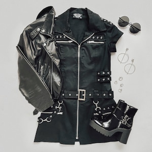 May include: A black short-sleeved dress with a full-length front zipper, silver-tone eyelets, buckles, and metal rings. The outfit includes a black faux leather jacket, black platform ankle boots with buckles, round sunglasses, and silver hoop earrings.