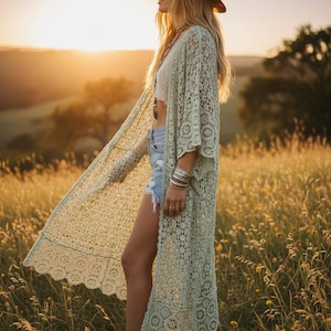 May include: A long, light green crochet duster cardigan featuring an intricate floral lace pattern, wide three-quarter length sleeves, and a scalloped hemline. The garment is worn over a white crop top and light blue denim shorts with frayed edges.