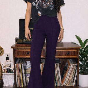 May include: Black Def Leppard graphic tee and high-waisted purple flared pants. The pants have a seam down the front and are paired with black studded shoes. A vintage record player and wooden cabinet are in the background.