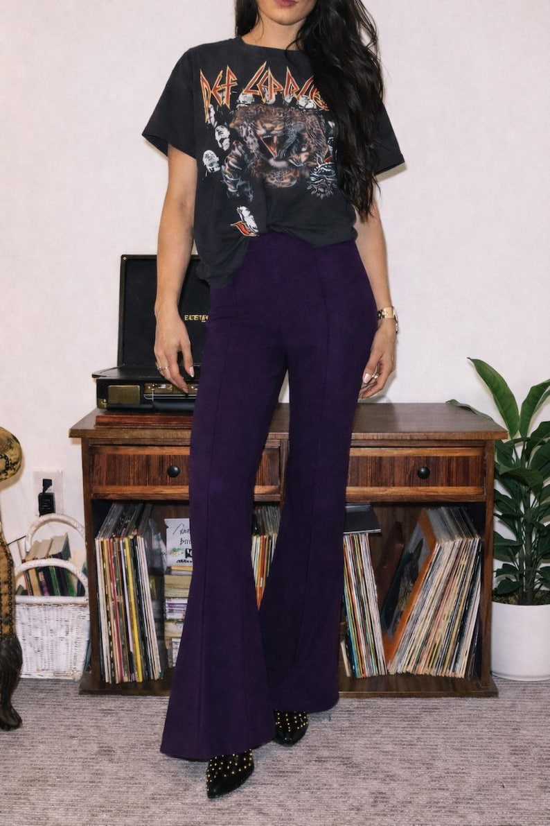 May include: Black Def Leppard graphic tee and high-waisted purple flared pants. The pants have a seam down the front and are paired with black studded shoes. A vintage record player and wooden cabinet are in the background.