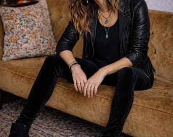 May include: A woman wearing a black leather jacket with fringe detail, black velvet pants, and black ankle boots. She is seated on a brown velvet sofa. Accessories include necklaces and rings. The background includes a lava lamp and a patterned pillow.