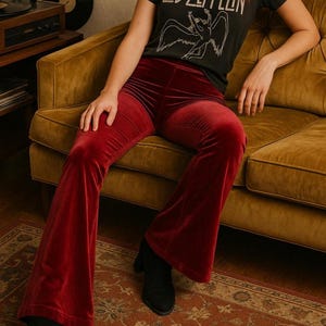 May include: A person wearing a black t-shirt with the Led Zeppelin logo and red velvet flared pants. The pants have a high waist and a wide leg. The person is sitting on a mustard-colored couch.