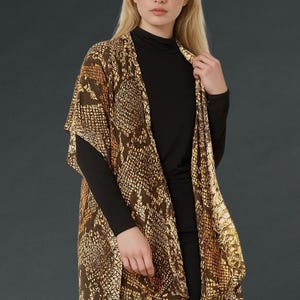 Puede incluir: Un cárdigan tipo kimono con estampado de piel de serpiente marrón y dorado. El cárdigan abierto se lleva sobre una camiseta negra de manga larga y pantalones negros. La modelo tiene el pelo rubio y está sobre un fondo gris oscuro.
