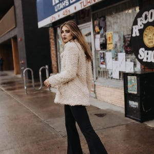 May include: A woman wearing a cream-colored, textured coat and black flared pants walks on a wet sidewalk. The background features a storefront with the sign "Randy's Records Vinyl Turntables" and a vintage record shop sign.