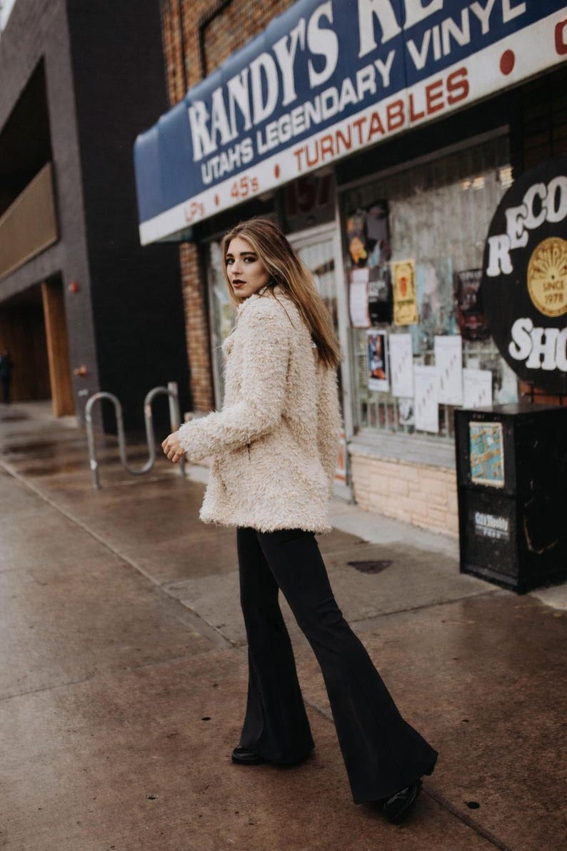 May include: A woman wearing a cream-colored, textured coat and black flared pants walks on a wet sidewalk. The background features a storefront with the sign "Randy's Records Vinyl Turntables" and a vintage record shop sign.