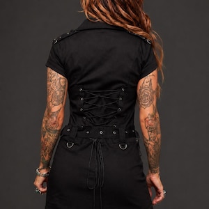 May include: Black short-sleeved dress with a corset-style back and a skirt. The dress features a lace-up back, D-ring details, and a short skirt. The model has tattoos on their arms and legs. The dress is a dark, gothic style.