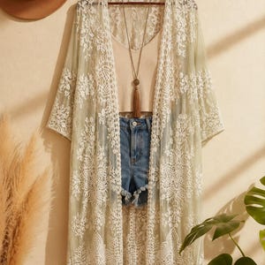 May include: A sage green open-front duster cardigan featuring intricate white floral lace patterns, worn over a beige ribbed tank top and blue denim shorts with frayed hems, accessorized with a long necklace featuring a brown tassel pendant.