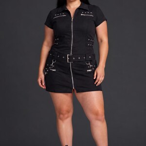 May include: A black short-sleeved mini dress featuring a full-length silver front zipper, a collared neckline, and a matching belt. The garment includes multiple silver-toned metal zippers, eyelets, and buckle details across the chest and skirt.